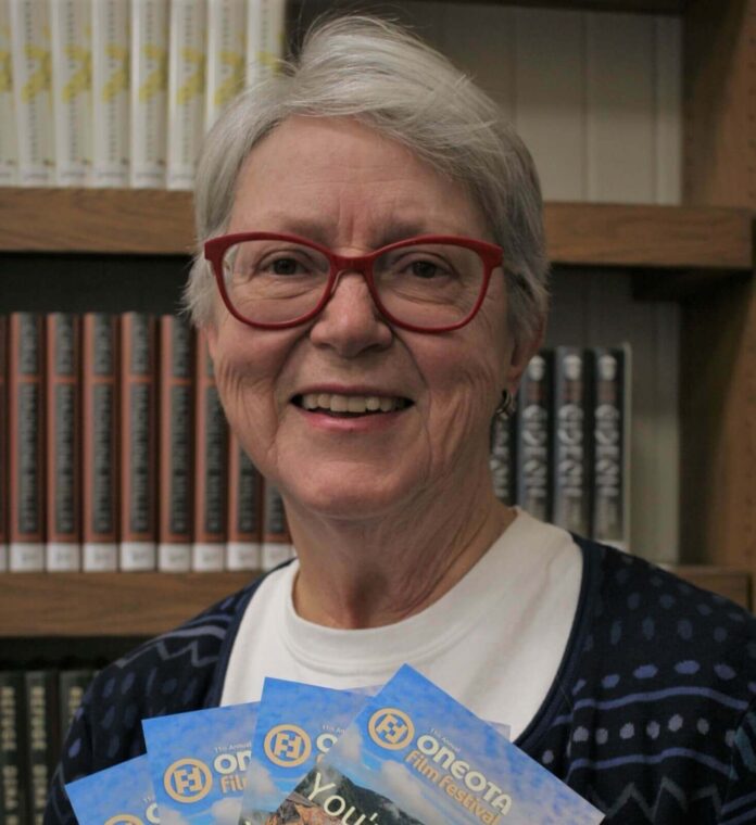 A smiling older woman with short gray hair and red glasses stands in front of a bookshelf. She holds several Oneota Film Festival brochures. She is wearing a patterned blue cardigan over a white shirt. Books are visible on the shelf in the background.