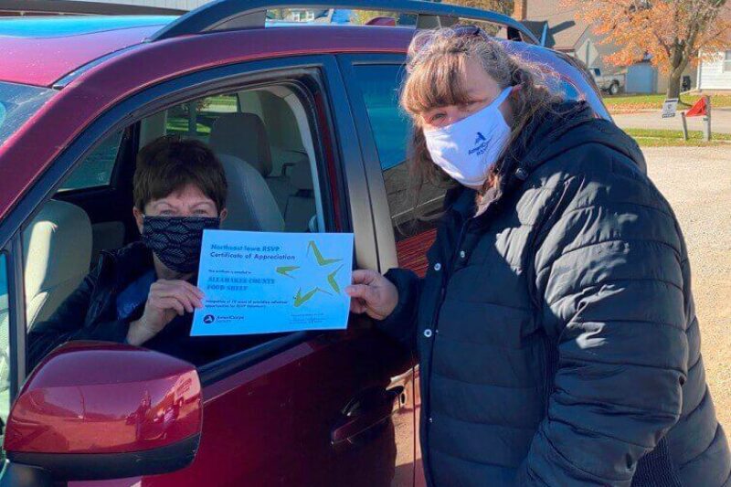 A person in a face mask sits in a red car holding a certificate. Another masked person stands outside the car door, smiling and looking at the camera. It appears to be a sunny day with autumn foliage in the background.