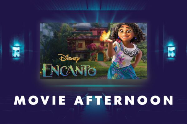 A digital graphic promoting a "Movie Afternoon" event features a dark blue theater setting with rows of seats and glowing blue wall lights. The central focus is a large movie screen displaying an image from Disney's Encanto, showing Mirabel Madrigal, a young girl with curly hair and glasses, wearing a colorful embroidered blouse and skirt, holding a glowing yellow butterfly. Behind her is the magical Madrigal family house. The Encanto logo appears in vibrant, multicolored text, with "Disney" written above it. Below the screen, "MOVIE AFTERNOON" is displayed in bold, white, all-capital letters. The image conveys a warm and inviting atmosphere for a film screening.